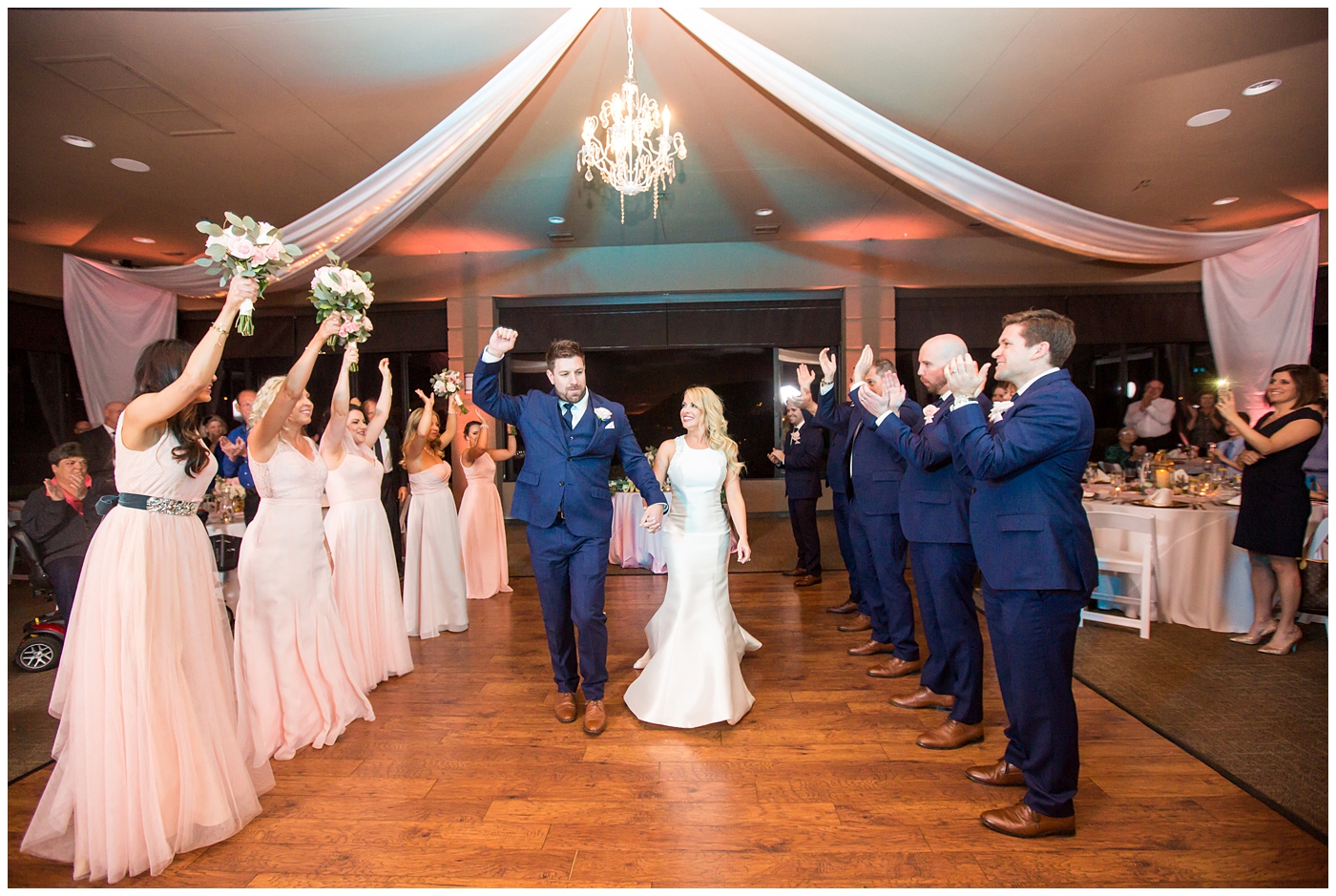 gorgeous bride with side swept hair in racerback pronovias dress and blush pink, white rose and eucalyptus greenery wedding bouquet with groom in navy suit and tie grand entrance at wedding reception in ballroom