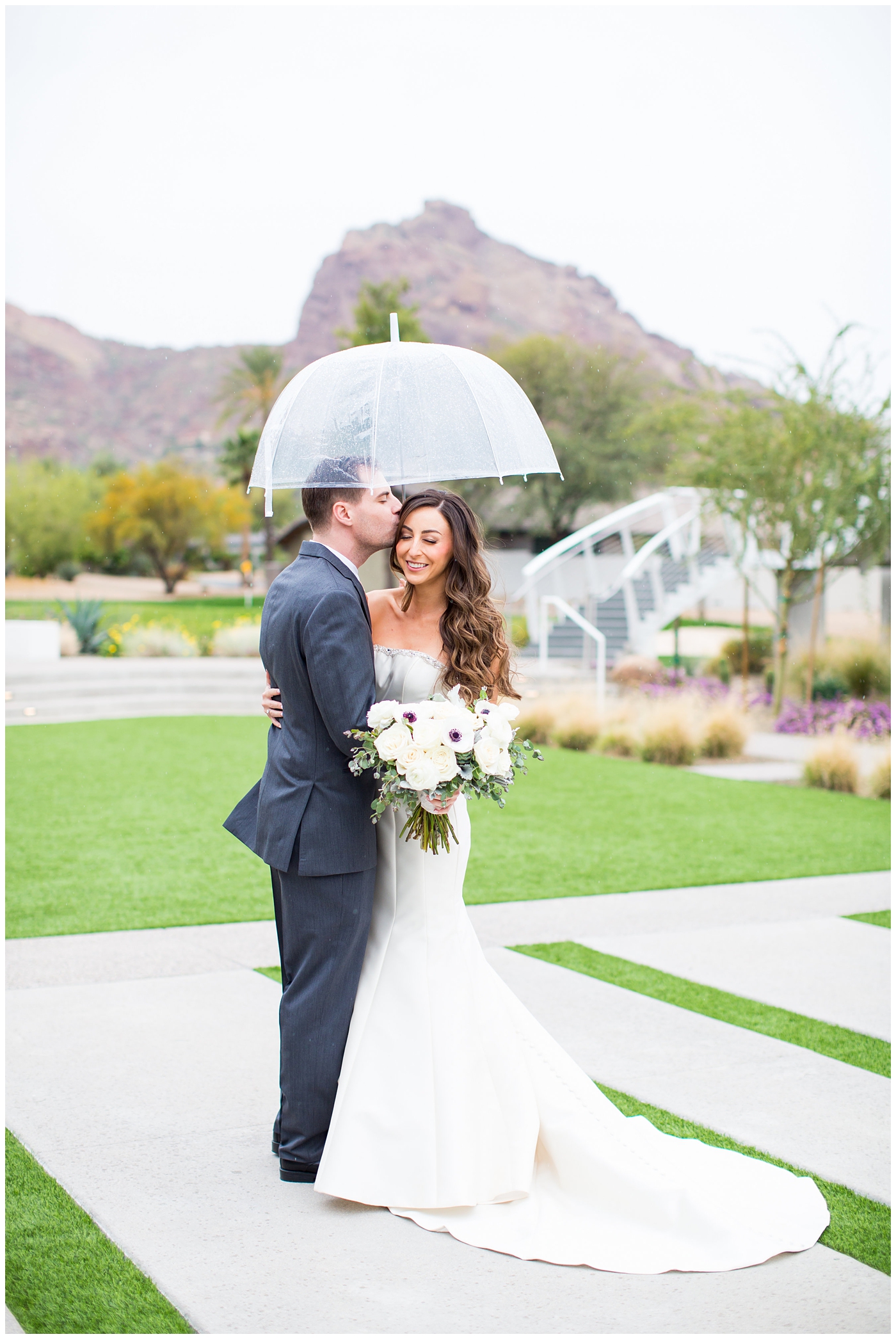 brunette bride getting into white strapless wedding dress with beaded top with White Anemones, roses, succulents, and greenery wedding bridal bouquet and groom in gray suit with gold tie wedding day portrait in the rain under clear umbrella
