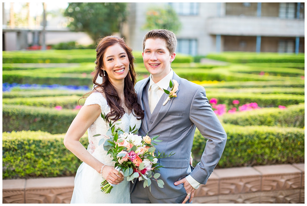 bride in matthew christopher wedding dress with cap sleeves with white, pink and orange ranunculus flowers and greenery bouquet with groom in light gray suit with tie wedding day portrait at Arizona Biltmore garden