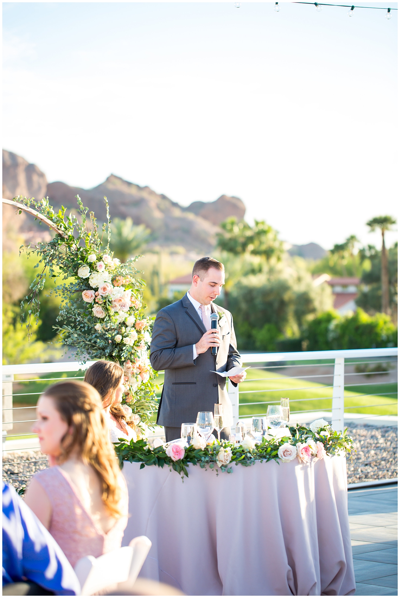 groom giving speech with unique circle arch for outdoor rooftop wedding reception with wildflowers in blush and white with greenery with mountain in background